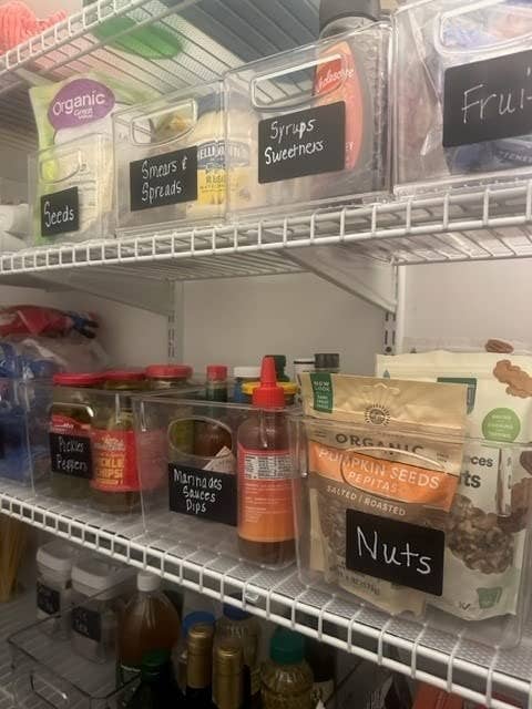 Organized pantry shelves with labeled bins for seeds, spreads, and syrups; various condiments in front