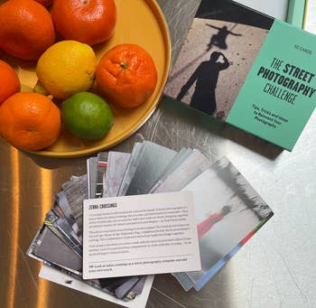 A selection of printed photographs laid on a surface with fruits on a plate nearby, and a book titled 'The Street Photography Challenge'