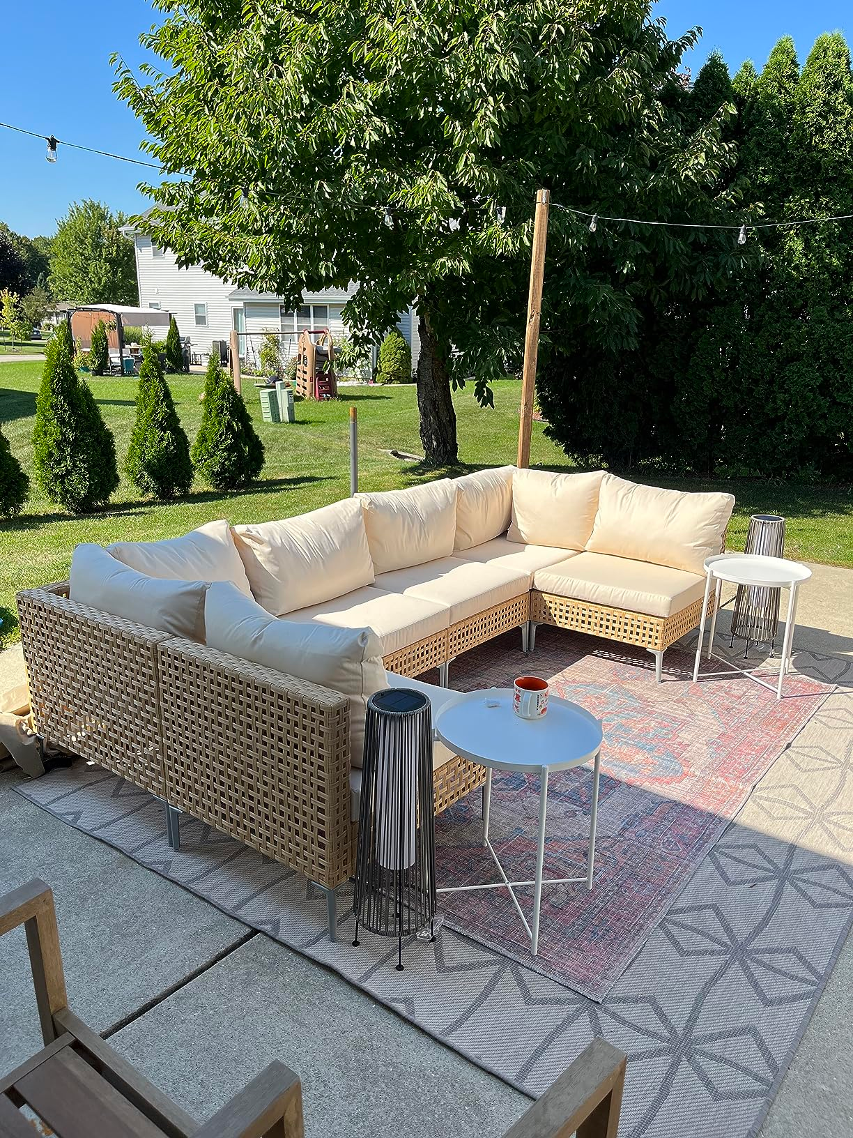 Outdoor wicker sectional with beige cushions, acceptable   connected  a patterned rug, surrounded by greenery. Two broadside  tables clasp  a candle and a lantern