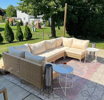 Outdoor wicker sectional with beige cushions, acceptable connected a patterned rug, surrounded by greenery. Two broadside tables clasp a candle and a lantern