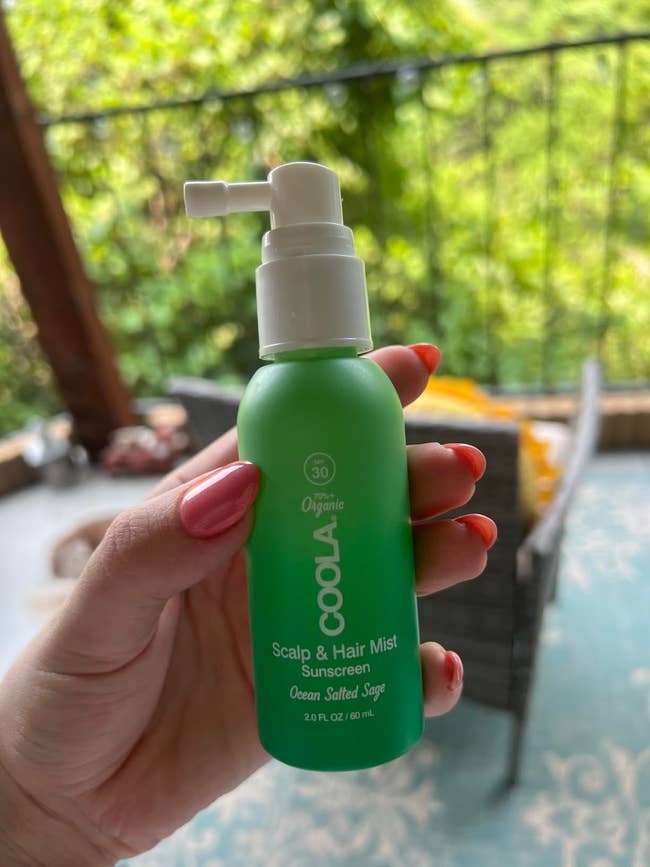 A manus holds a greenish vessel of COOLA Scalp & Hair Mist Sunscreen, labeled Ocean Salted Sage, against a patio background