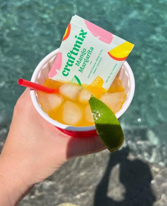 Person holding a cup with ice and a mango margarita drink mix packet