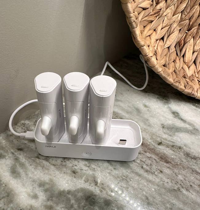 Three achromatic  portable chargers connected  a charging dock, placed connected  a marble aboveground  adjacent  to a woven basket