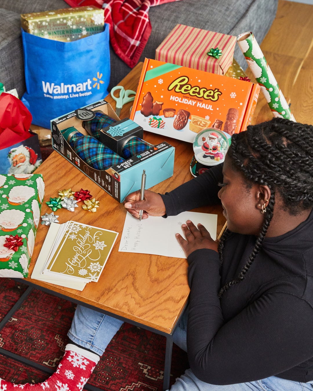 Elise writes in holiday card on table next to Reese's holiday haul candies, Santa snow globe, Walmart bag, and plaid 3-piece bottle set