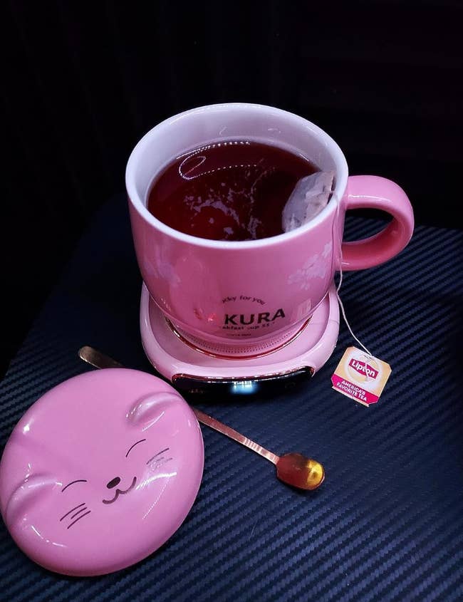 A pink mug of tea on top of a matching pink warmer with a cat face-shaped mug topper on the side 