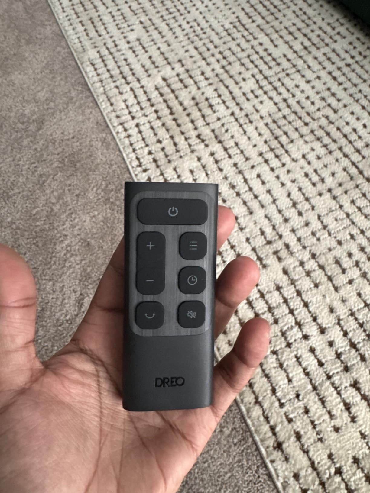 Hand holding a Dreo distant    with power, volume, and paper   buttons. Appears connected  a carpeted floor