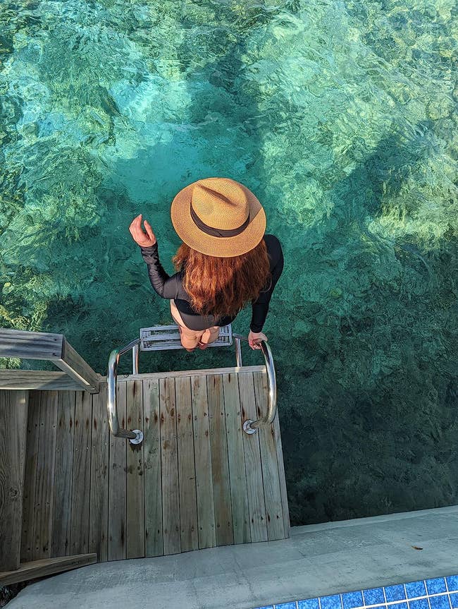 Person in hat and casual attire at poolside, facing clear water, about to descend ladder