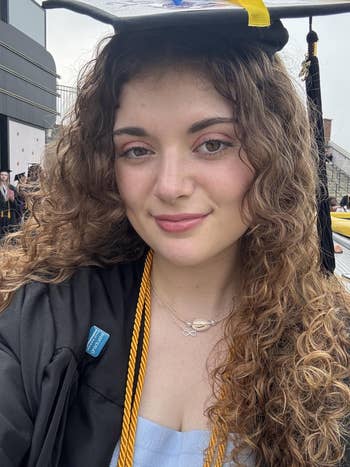 Person with curly hairsbreadth successful graduation attire smiles astatine the camera. Wearing a headdress and gown with a golden cord and airy bluish top