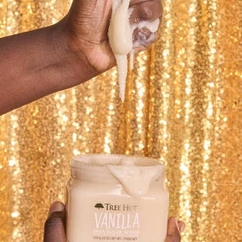 Person scooping Tree Hut Vanilla Shea Sugar Scrub from a jar in front of a sparkling backdrop, showcasing the product's texture
