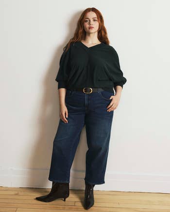 Person stands indoors wearing a relaxed V-neck sweater, belted jeans, and ankle boots, suitable for casual buying  attire
