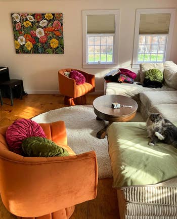 A cozy living room with a cat on the couch, adjacent armchairs, a coffee table, and a floral painting above