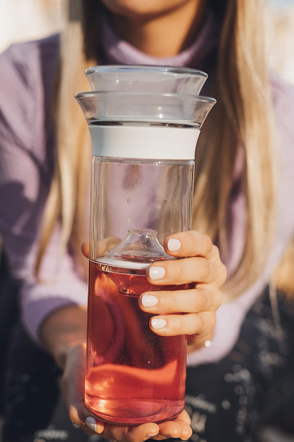 Person holding a glass infuser bottle filled with a beverage. The bottle has a unique design with an inner infuser chamber and a white band around the top
