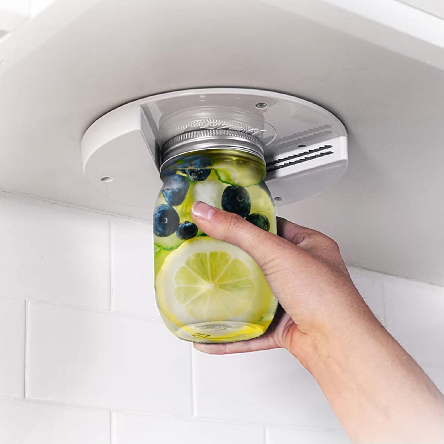 A hand is using an under-cabinet jar opener to open a jar filled with lemon slices and blueberries