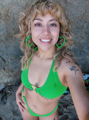 Woman in green bikini, hoop earrings, with a rock background, smiling at the camera