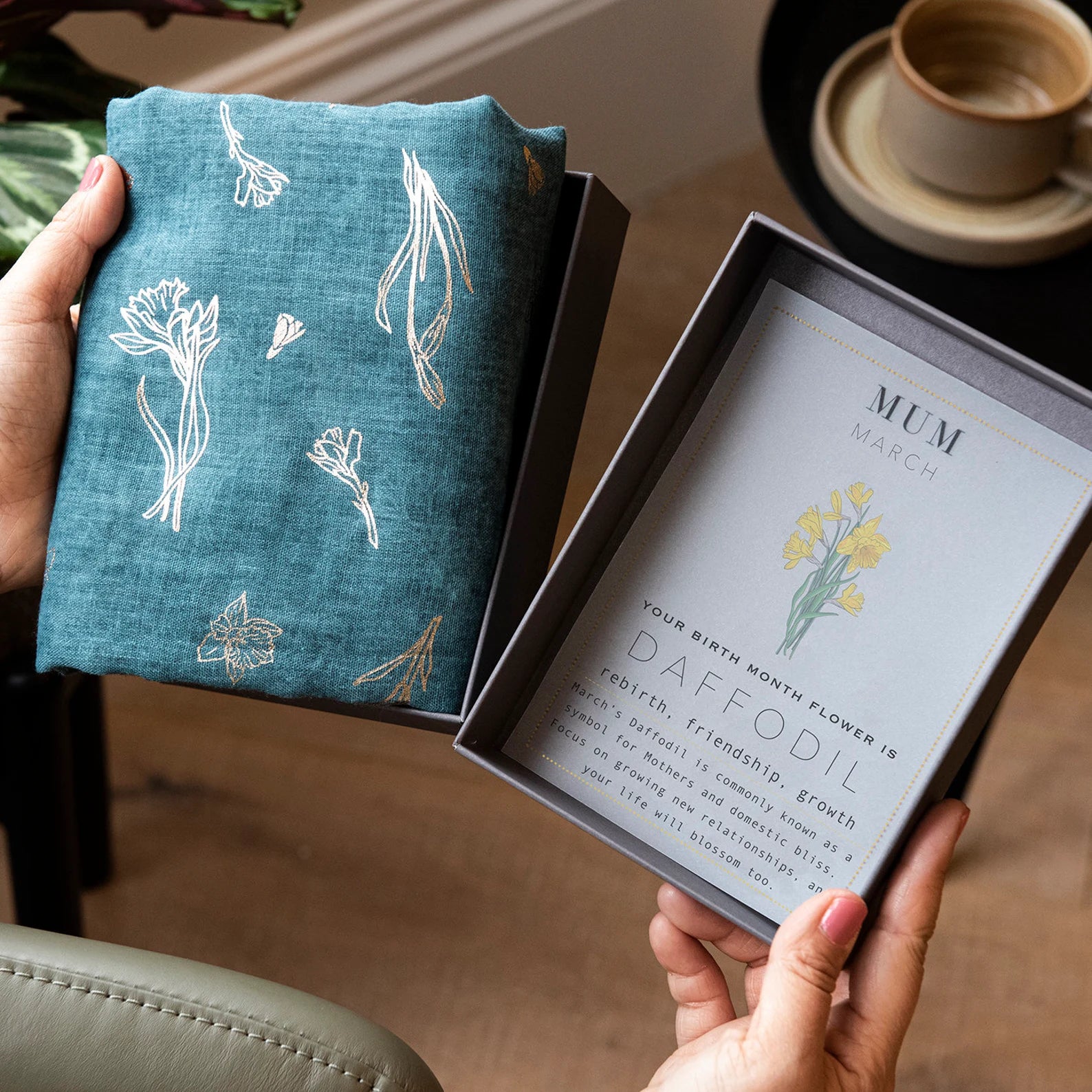 person holding an open box with a blue and gold birth month flower scarf on the left side, and an info card about the