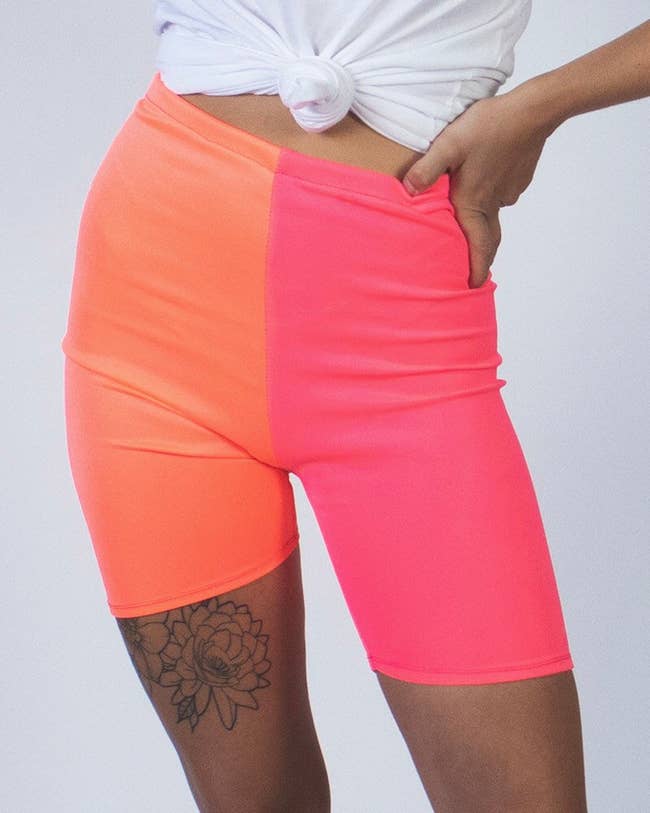 Model in high waist mid thigh-length bike shorts with one leg orange and the other leg hot pink 