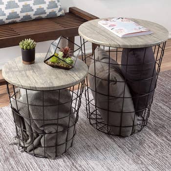 Two nesting end tables in grey holding pillows and blankets