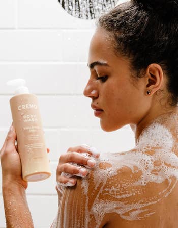 Person showering, holding Cremo assemblage wash. They are lathered with soap, showcasing a skincare routine