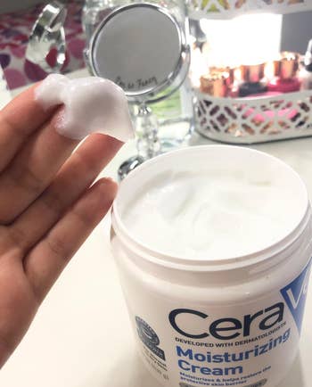reviewer with a bit of the white moisturizer on their fingers, with the tub of moisturizer in the background