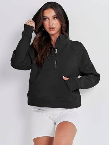 Woman in black hoodie and white shorts standing, hand on head, fashion pose for shopping article
