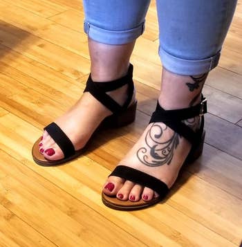 Person wearing black sandals with ankle straps and red nail polish, displaying a tattoo on their leg. Cropped jeans on a wooden floor