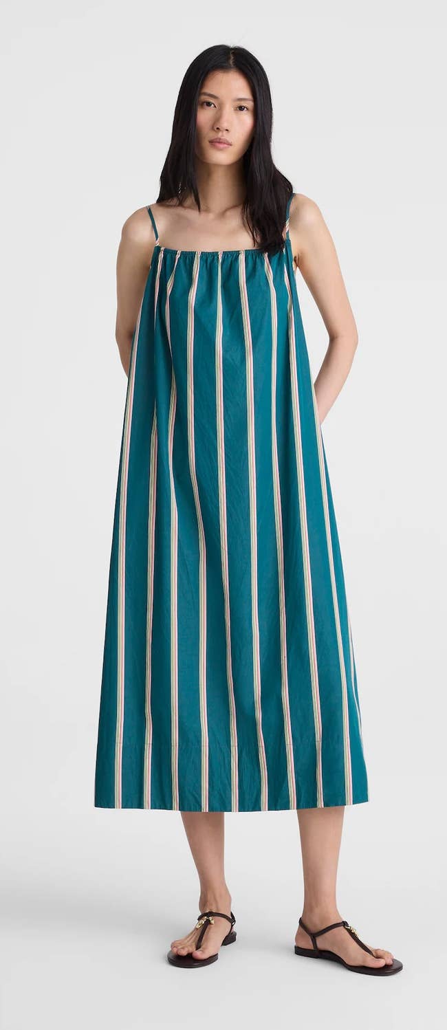 Model in a loose, green striped maxi dress and sandals stands against a plain background