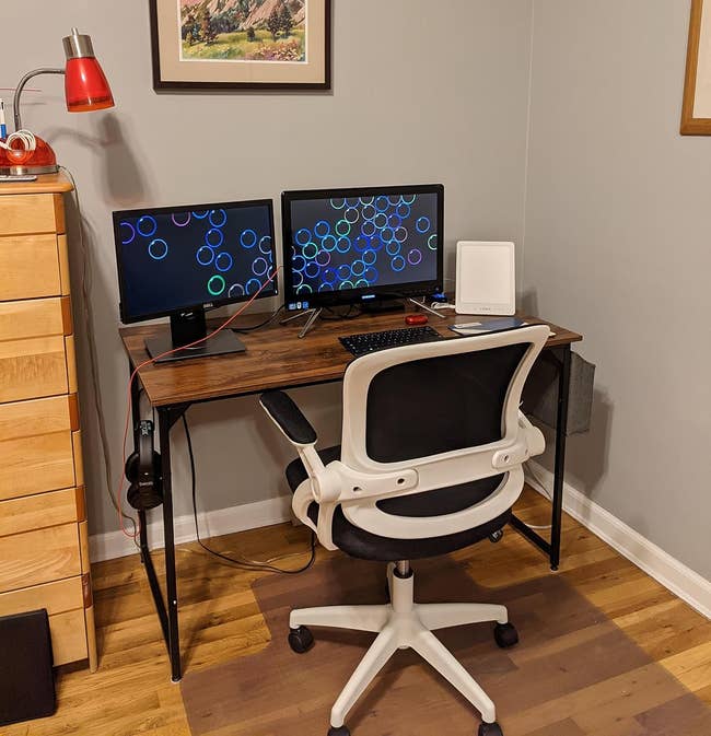 Home office setup with a desk, chair, dual monitors displaying a pattern, a lamp, and framed wall art. Featured in a shopping article