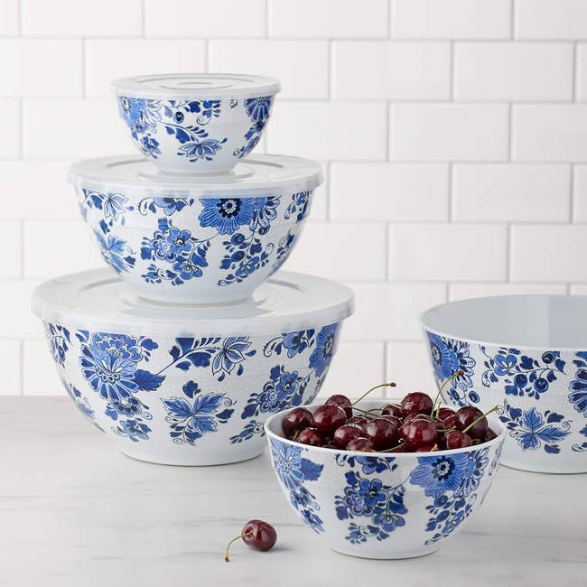 Stacked bluish  floral bowls with lids adjacent  to a vessel  of cherries connected  a room  counter, acceptable   against a tiled backsplash