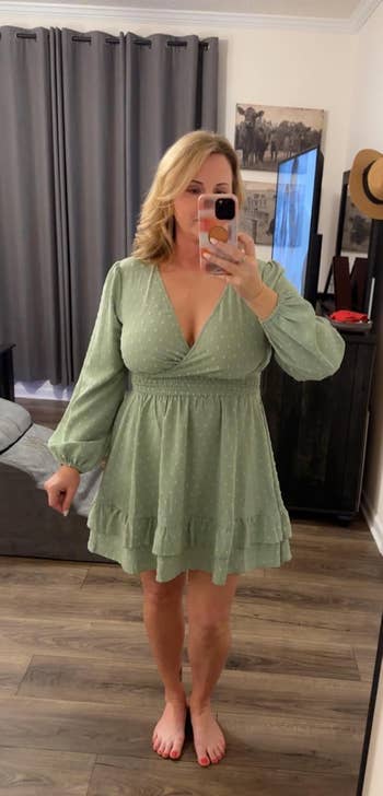 A person takes a mirror selfie at home, wearing a flowy dress with long sleeves and a V-neckline. Room features gray curtains and modern decor