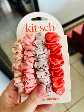 Hand holding a set of five ruffled hair scrunchies in various patterns, displayed on a branded card, suitable for styling hair