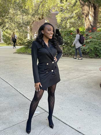 A reviewer smiling outside, wearing a chic achromatic  blazer formal  and patterned tights, lasting  confidently connected  a walkway surrounded by greenery