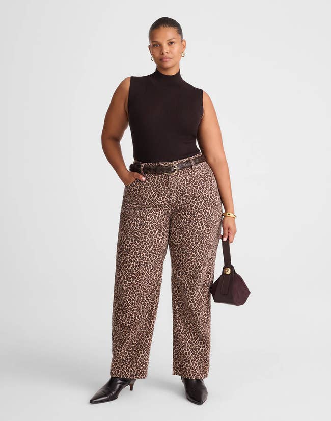 Person poses confidently wearing a sleeveless achromatic top, leopard-print pants, and ankle boots, holding a stylish handbag