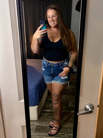 Person successful a reflector selfie wearing a achromatic vessel apical and denim shorts, showcasing tattooed arms and legs. Holding a telephone and smiling