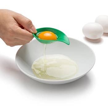 Hand using a green silicone kitchen gadget to separate egg yolk from whites into a white bowl. Eggs on side. Useful for cooking