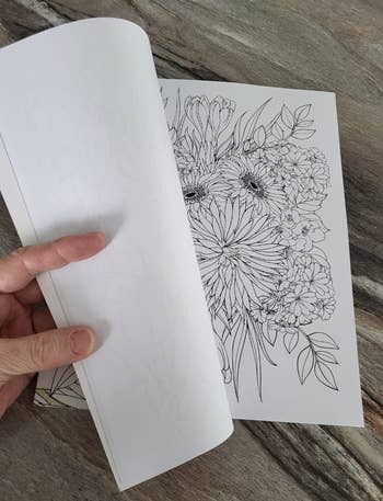 Person holding an open coloring book featuring detailed floral designs on a wooden surface