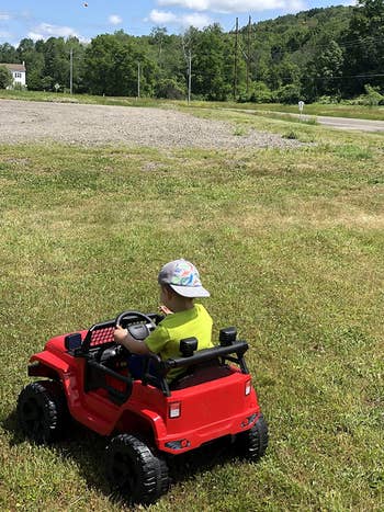 Child in a toy car wearing a bucket hat and casual clothing, outdoors on grass