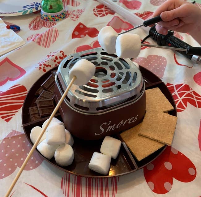 a reviewer photo of indoor s'mores maker with marshmallows on sticks being heated, chocolate squares and graham crackers ready for assembling