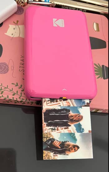 A portable printer prints a photograph  of 2  women outdoors, 1  holding a cup. The printer is connected  a pinkish  publication  with illustrations