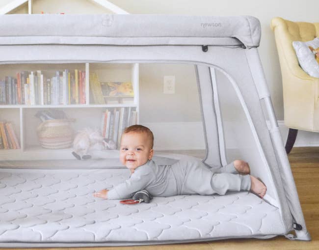 Baby smiling connected a padded mat wrong a mesh playpen, with a bookshelf and toys successful the background