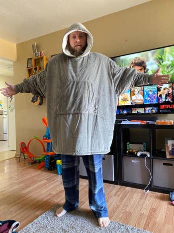 Person wearing an oversized, hooded gray sweatshirt and plaid pajama pants, standing in a living room with a TV in the background