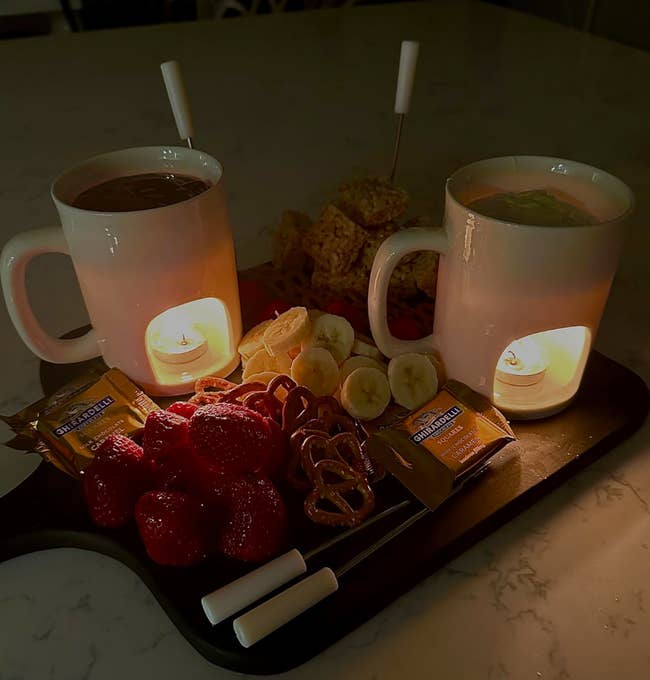 Two mugs with beverages, surrounded by strawberries, bananas, pretzels, and snack bars on a tray