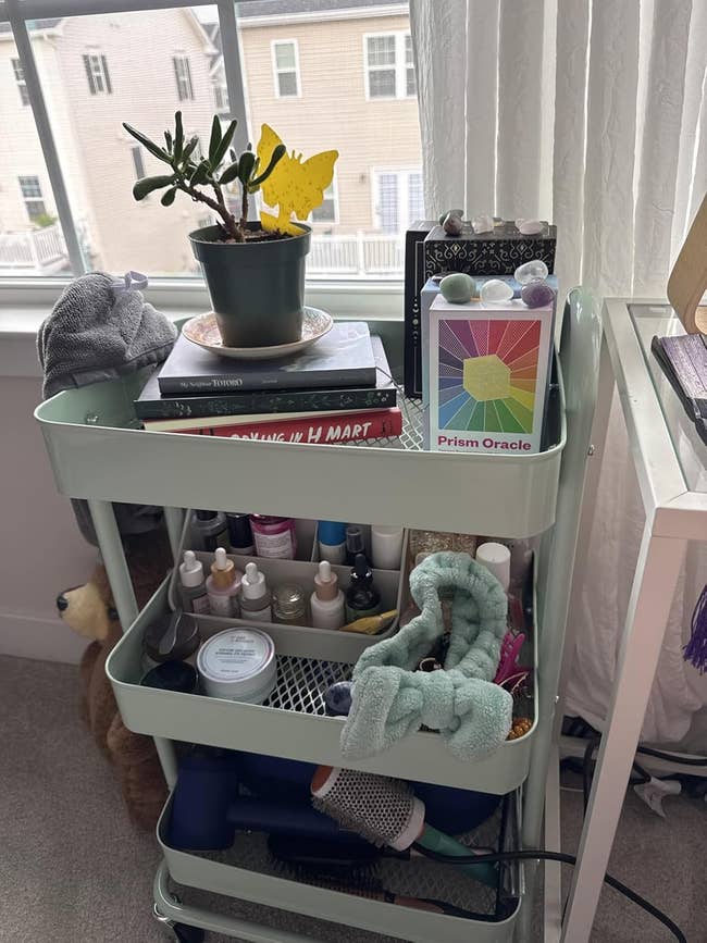 Rolling cart with skincare products, hairsbreadth tools, a plant, books, and a paper deck. Placed by a window, the cart organizes quality and self-care items