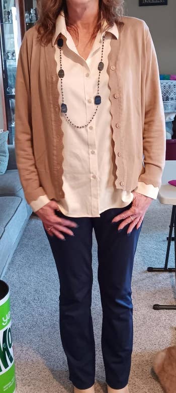 Person in a casual outfit with a beige cardigan, white blouse, blue pants, and a statement necklace, standing in a living room