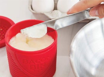 hand taking some solidified bacon grease out of the bacon bin with a knife