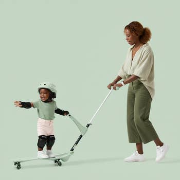 Toddler with helmet and knee pads on a scooter, guided by an adult wearing casual attire, in a shopping context