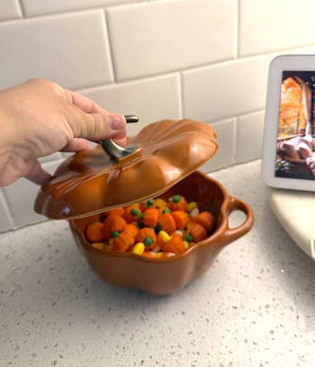 Hand lifting lid of pumpkin-shaped pot, revealing candy pumpkins inside, connected a room antagonistic with a tablet nearby