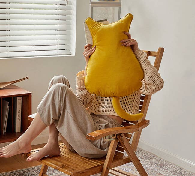 Model is sitting on a chair holding a mustard yellow cat shaped pillow