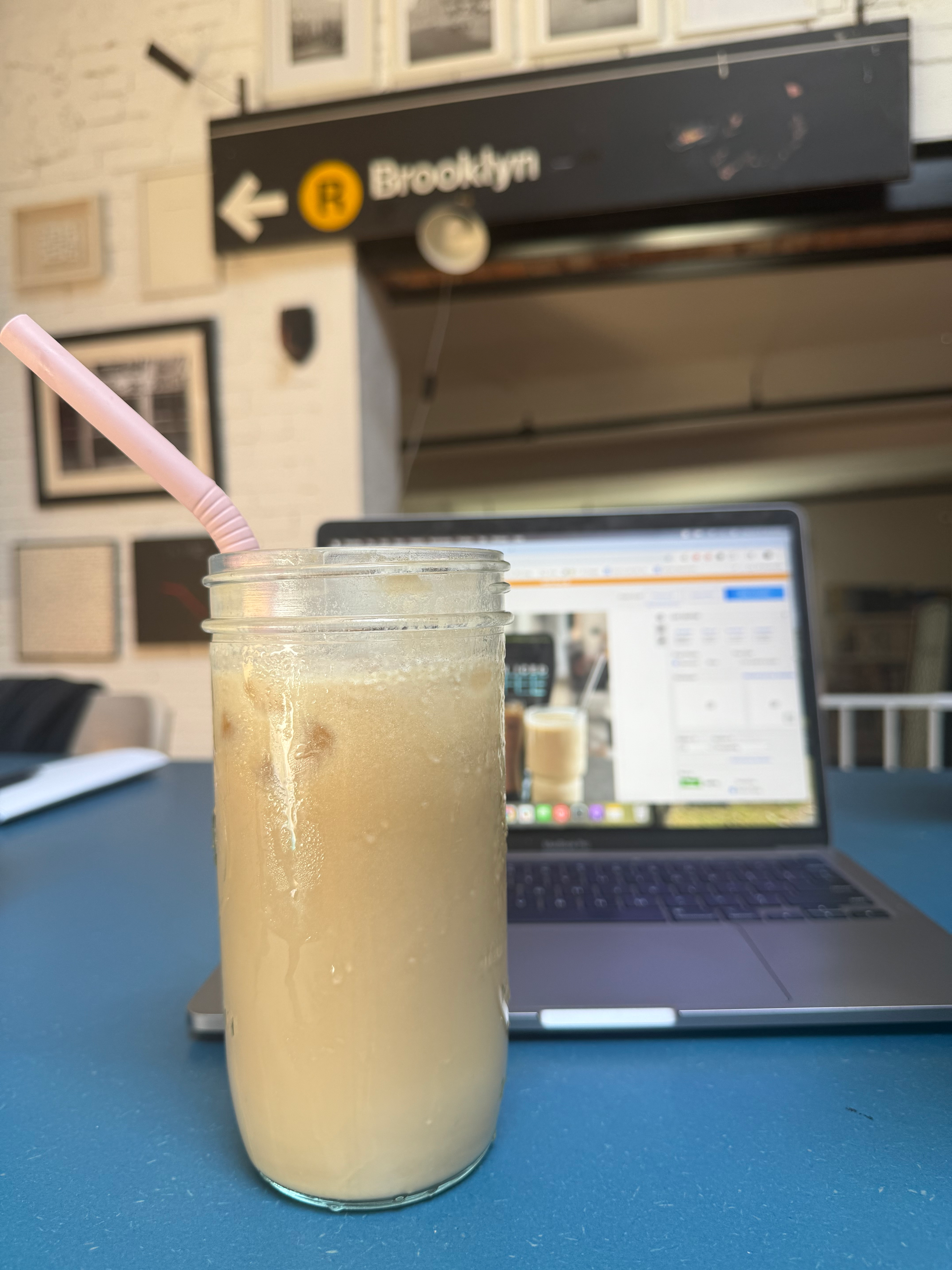 Es kopi dalam toples kaca dengan sedotan, diletakkan di atas meja di sebelah laptop terbuka di pengaturan kafe yang trendi