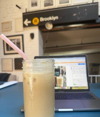 Es kopi dalam toples kaca dengan sedotan, diletakkan di atas meja di sebelah laptop terbuka di pengaturan kafe yang trendi