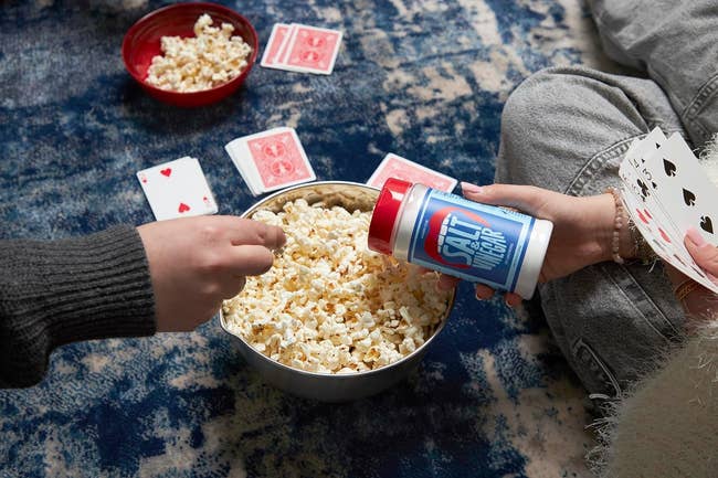 Two people playing cards and adding seasoning to popcorn on a floor